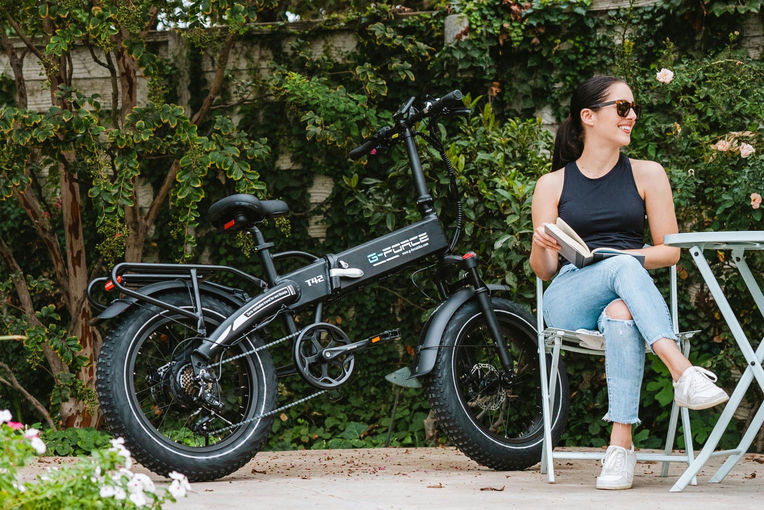 Woman in casual attire reading next to an electric bike on a sunny day.