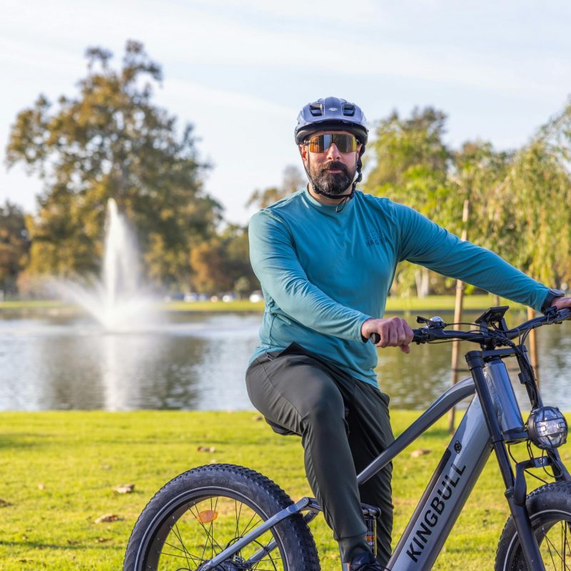 A man wearing a helmet rides a bicycle in a scenic park near a fountain.